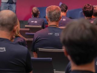 Crowd in an auditorium wearing navy blue CI&T T-shirts with AWS logos, seated on dark blue chairs against a red backdrop.