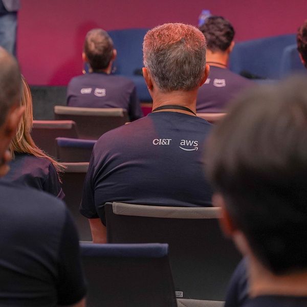 Crowd in an auditorium wearing navy blue CI&T T-shirts with AWS logos, seated on dark blue chairs against a red backdrop.