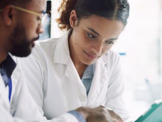 Close-up of two pharmacists. The man on the left has glasses and a beard, while the woman on the right has curly hair pulled back. Both are looking down at a tablet.