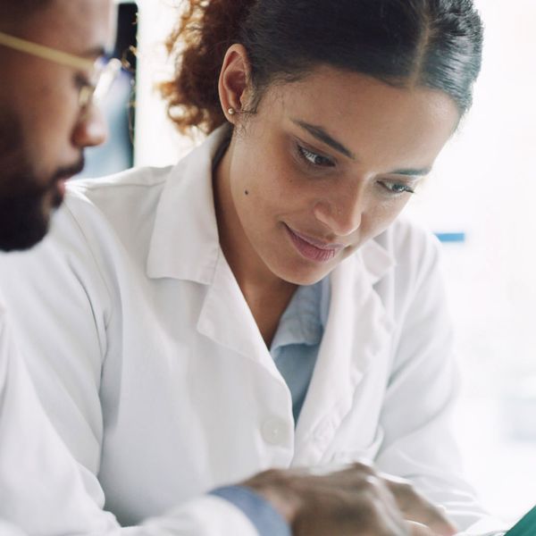 Close-up of two pharmacists. The man on the left has glasses and a beard, while the woman on the right has curly hair pulled back. Both are looking down at a tablet.