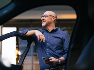 A man stands next to a car, casually leaning against the open door while holding a smartphone, showcasing a modern urban setting.
