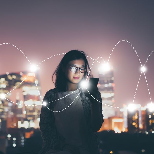 A woman in front of a cityscape at night looking at her smartphone